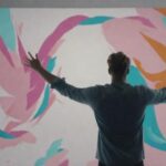 A masculine presenting designer waves their hands in front of an abstract colorful canvas.
