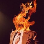 Cropped image of a man in a brown suit and tie whose face is engulfed in fire from the Late Night With The Devil official movie poster.